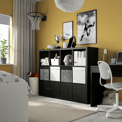 Estantería negra KALLAX junto al escritorio, con papeleras blancas para material deportivo. El diseño moderno destaca sobre la pared amarilla, con un aro de baloncesto encima.