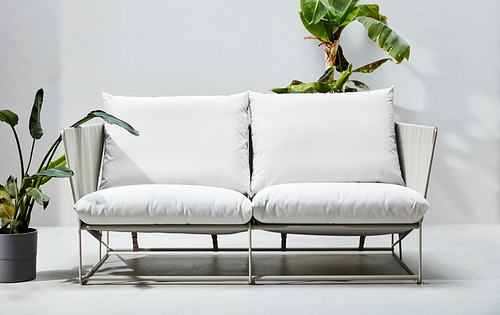 Moderno sofá de dos plazas blanco con estructura de metal, cojines de felpa y decoración de plantas frondosas.