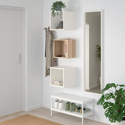 Pequeños estantes EKET en la pared, blancos y de madera, que almacenan libros y decoración, con un gran espejo al lado, debajo de un gancho para la ropa.