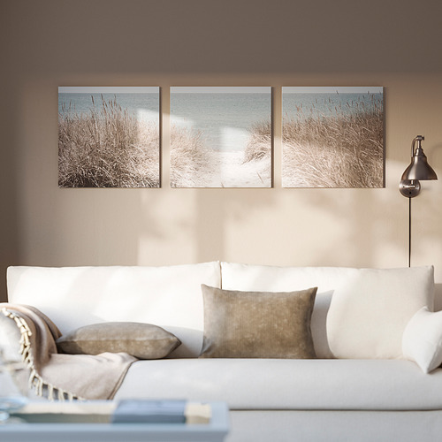Three beach photos in white frames on brown wall. White sofa with pillows, blanket, and silver lamp beside it.