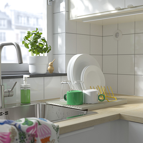 Fregadero de cocina con escurridor de platos RINNIG que sostiene platos, tazas y cubiertos. Jabón verde para platos y planta cercana.