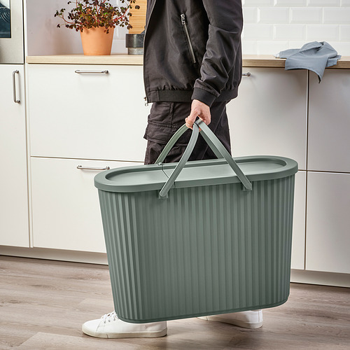 Persona sosteniendo un cubo de basura verde con el asa levantada, listo para el transporte. La tapa oculta el contenido, se adapta a espacios estrechos.