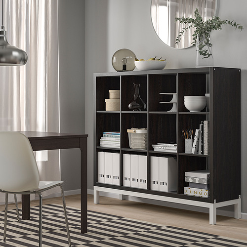 La estantería negra KALLAX contiene varios artículos, de pie junto a un escritorio y una silla, con libros y decoración cuidadosamente organizados.