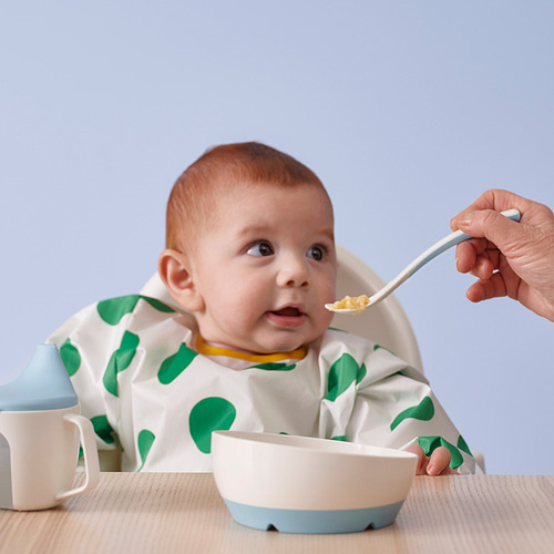 El bebé se sienta en una silla alta, con un babero de lunares verdes, y es alimentado con cuchara de un tazón con borde azul y una cuchara de mango largo.