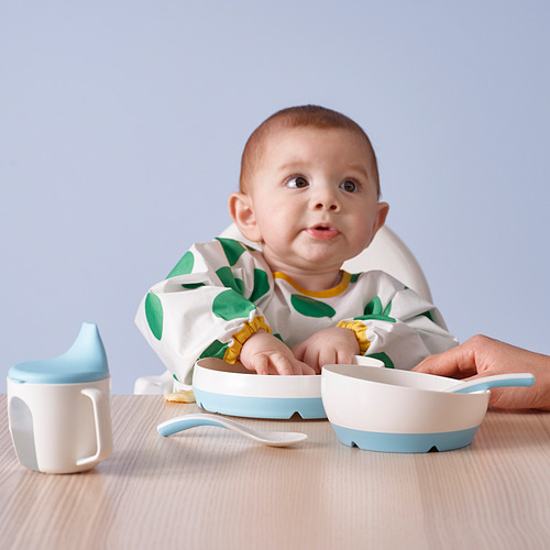 El bebé con babero de lunares verdes se sienta a la mesa con el juego de cucharas BÖRJA: una cuchara para alimentarse solo, otra para usar en tarro.