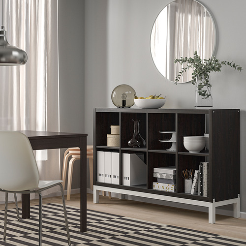Habitación moderna con estante KALLAX marrón oscuro, cuenco blanco, jarrón, libros, espejo, silla beige, alfombra a rayas. El estante es grande, cabe debajo del espejo, al lado de la silla.