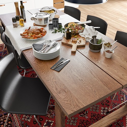 Mesa de comedor moderna de roble con sillas negras, preparada para una comida. Fíjate en la veta rústica, el mantel blanco y varios platos y utensilios.