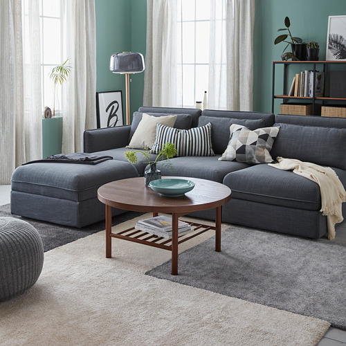 Sala de estar moderna con un gran sofá gris en forma de L, varios cojines, una mesa de centro circular de madera y una alfombra beige sobre una alfombra gris.