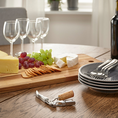 Mesa de madera con queso, uvas, galletas, copas de vino, platos, tenedores, un sacacorchos y una botella, lista para una comida.