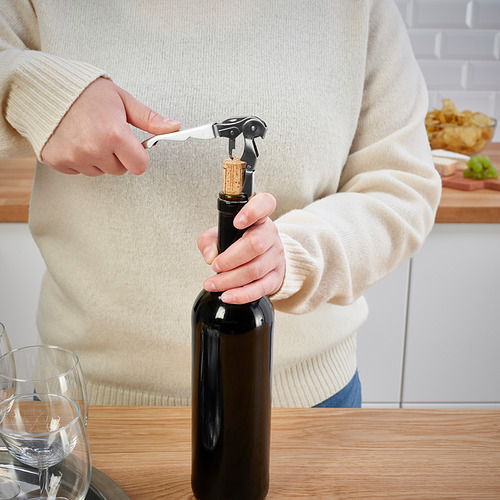 La persona abre una botella de vino con un sacacorchos. Escena sobre mesa de madera con vasos.
