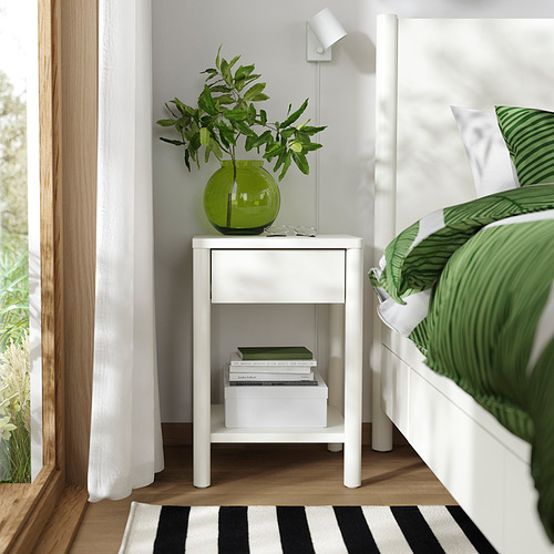 Mesita de noche de color blanquecino con libros y jarrón verde cerca de la cama.