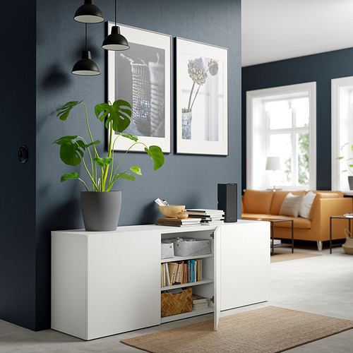 Sala de estar moderna con mueble blanco BESTÅ, decorado con libros, fotos, lámparas, y una gran planta, sofá naranja, pared azul marino.