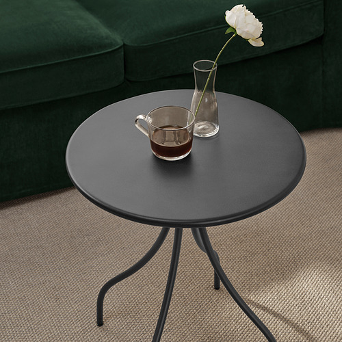 Mesa auxiliar redonda negra de 3 patas, bebida y flor, sobre alfombra beige, junto a sofá verde.