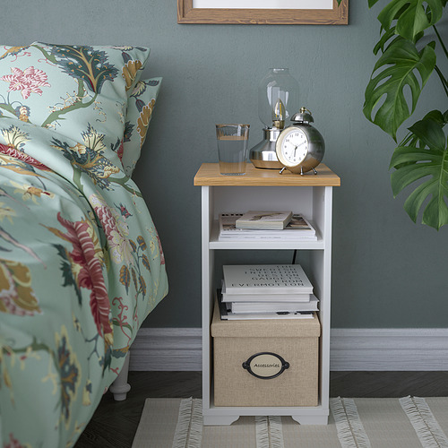 Una mesa auxiliar SKRUVBY con tapa blanca y de madera, con libros, un reloj y una lámpara. Junto a una cama con ropa de cama floral.