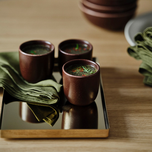 Tres tazas de cerámica marrón sostienen hierbas verdes en una bandeja de madera, con una servilleta de lino verde.