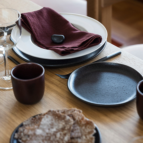 Un plato hondo blanco STOCKHOLM 2025 con borde plano, engastado con una servilleta granate y piedra, colocado sobre una mesa de madera con otros platos y vasos.