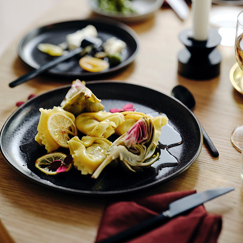 Platos negros mate con alcachofa y rodajas de limón, espaciados sobre una mesa de madera con cuchillos y vasos.