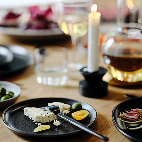 Plato oscuro con queso, limón, aceitunas; vela, vaso, cuenco al lado.
