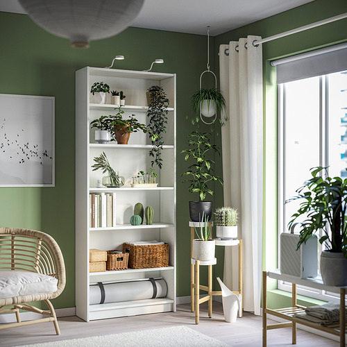 Una acogedora habitación verde con una estantería BILLY, llena de plantas y libros, acompañada de una silla de mimbre y cortinas.