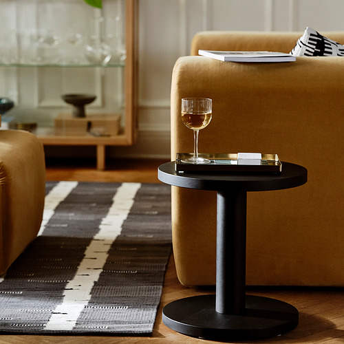 Mesa auxiliar negra con tapa redonda, vaso y posavasos. Al lado del sofá sobre alfombra. Diseño escandinavo.