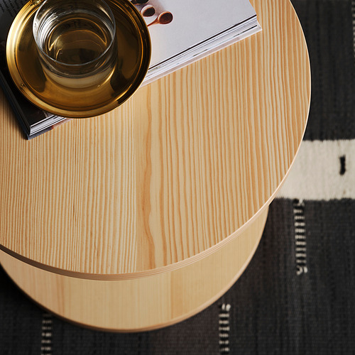 Vista superior de la mesa auxiliar minimalista de madera clara STOCKHOLM 2025 con taza de latón, libro negro, sobre alfombra estampada.