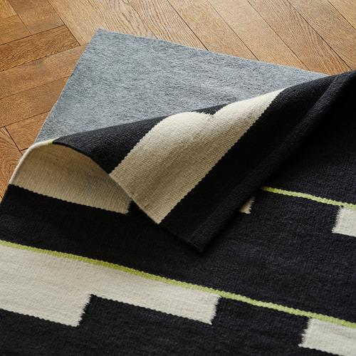 Esta imagen muestra una alfombra de lana reversible tejida a mano con un patrón geométrico en negro, blanco y gris. La alfombra se extiende sobre un suelo de madera.