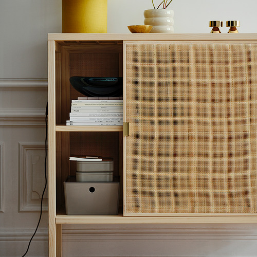 Un armario de madera parcialmente abierto con puertas de ratán, libros cuidadosamente apilados y cajas de almacenamiento. Las asas de latón acentúan su elegante diseño.