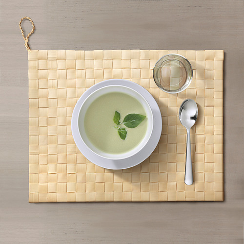 Un plato de sopa verde sobre un mantel individual rectangular de hojas de palmira tejido a mano con una cuchara y un plato pequeño. La alfombrilla tiene un lazo para colgar.