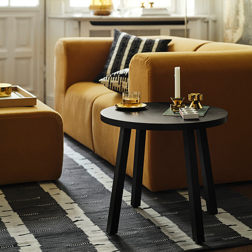 Sala de estar moderna con sofá seccional mostaza, mesa auxiliar redonda negra y alfombra a rayas. Mesa, candelabros de oro y un vaso.