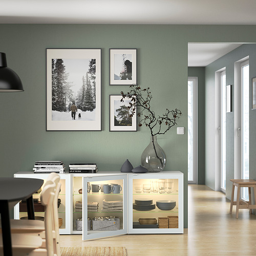 Moderna zona de comedor con aparador BESTÅ con puertas de cristal, platos expuestos y decorado con obras de arte enmarcadas y un jarrón de cristal con ramas.