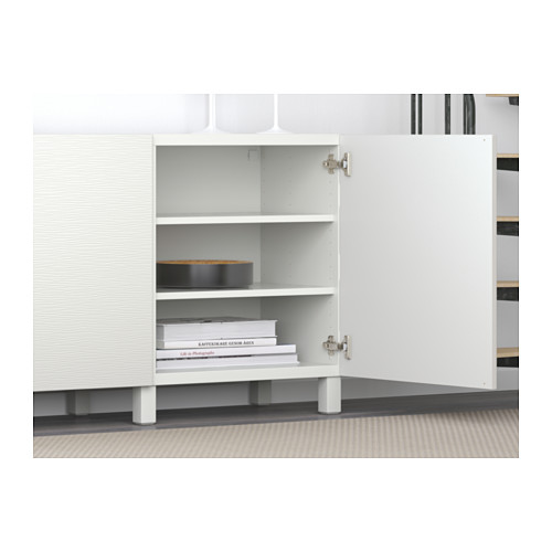 Mueble de almacenamiento blanco BESTÅ con estantes, una puerta abierta que revela libros y decoración. Bisagras de cierre suave visibles.
