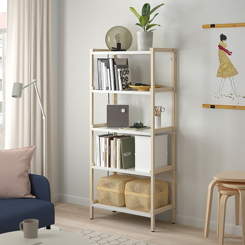 La estantería de madera clara EKENABBEN contiene libros, cestas de decoración y almacenamiento, colocadas entre el sofá y la pared, rematadas con una lámpara.