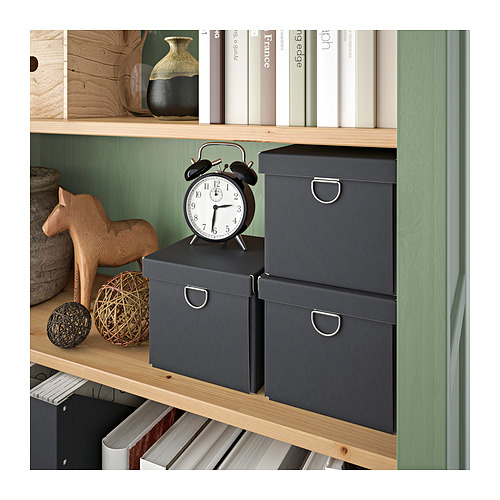 Una librería HEMNES en un relajante tono gris verdoso con estantes de madera teñida. Exhibe artículos decorativos, libros y cajas de almacenamiento en los estantes.