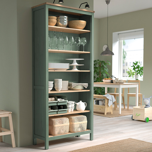 Una estantería alta HEMNES verde con estantes de madera. Se encuentra contra una pared verde, sosteniendo varios utensilios de cocina y rodeado de muebles de madera.