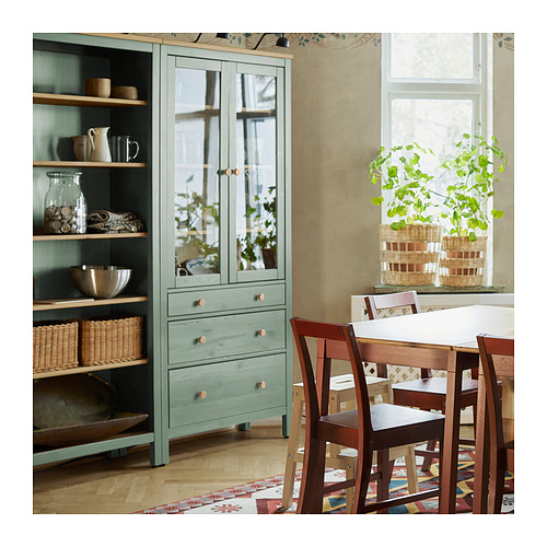 Acogedora zona de comedor con armario HEMNES verde, jarrones y frascos. Sillas marrones alrededor de una mesa blanca junto a una ventana frondosa.