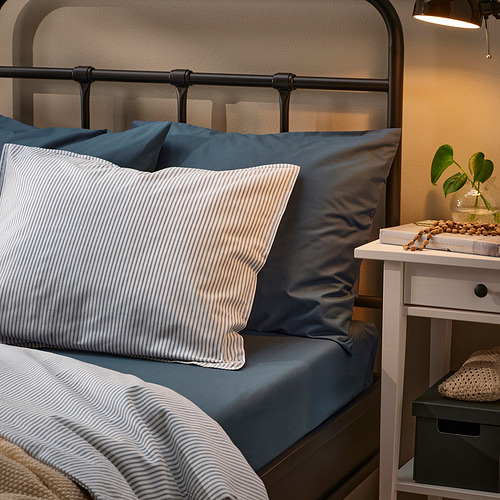 Acogedora configuración de cama con sábanas DVALA azules, almohadas a rayas y una cabecera de metal negro; Pequeña planta y libros en mesita de noche.