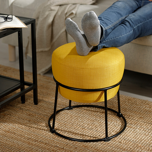 Persona reclinada con los pies en alto sobre un puf de EMMISLÖV de color amarillo brillante, que muestra su uso como reposapiés.