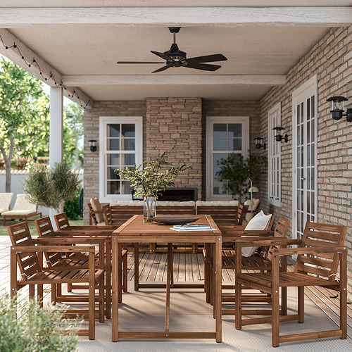 Juego de comedor al aire libre con mesa, sillas y ventilador. Telón de fondo de edificio de ladrillo con plantas. Luminoso y acogedor.