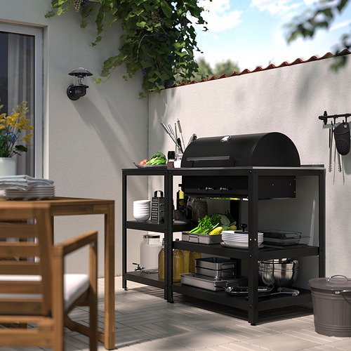 Estación negra de barbacoa GRILLSKÄR en el patio con estanterías, herramientas y zona de preparación de alimentos, rodeada de mobiliario de comedor.