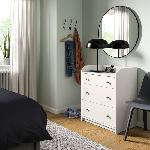 Dormitorio minimalista con cofre HAUGA blanco, espejo redondo, lámpara negra y ropa de cama oscura.