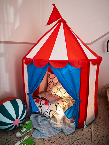 Carpa de juego para niños brillantes con forma de carpa de circo, parte superior a rayas rojas y blancas con paredes azules, acogedor en el interior con almohadas y una pequeña lámpara.