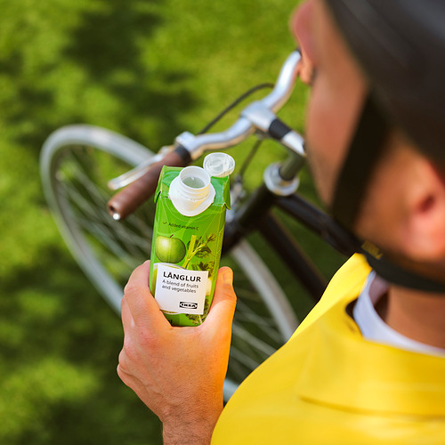 Una persona sostiene un batido verde de LÅNGLUR y muestra la etiqueta mientras está al aire libre en bicicleta.