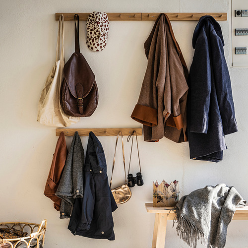 Un perchero de madera, HÖVOLM, con chaquetas, bolsos y accesorios. Tiene un acabado en roble natural y está montado en una pared.