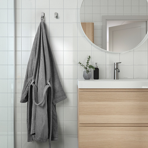 Albornoz gris con bolsillos cuelga de un gancho de pared junto al moderno lavabo del baño.