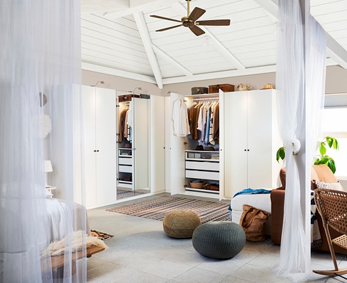 Dormitorio con armarios blancos, uno abierto mostrando ropa; Espejo adjunto, pisos de madera, alfombra beige, silla de mimbre, dos pufs redondos, ventilador de techo.