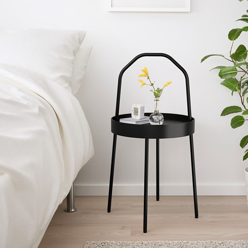 Mesita de noche moderna minimalista de metal negro al lado de la cama, sosteniendo un pequeño jarrón y un reloj despertador, junto a una gran planta verde.