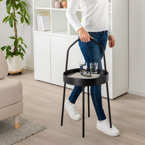 Persona sosteniendo una mesa auxiliar redonda portátil con asa. La mesa tiene capacidad para dos vasos y posavasos. La mesa tiene un marco de metal negro y una tapa de color claro.