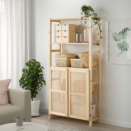 Estantería IVAR de madera clara con puertas de bambú en un espacio luminoso que contiene almacenamiento entre el sofá y la planta.