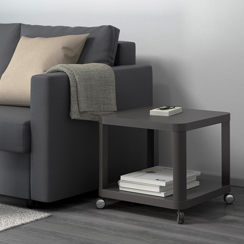 Sala de estar moderna con mesa auxiliar TINGBY gris junto al sofá, sosteniendo libros y revistas, con una manta en el reposabrazos.