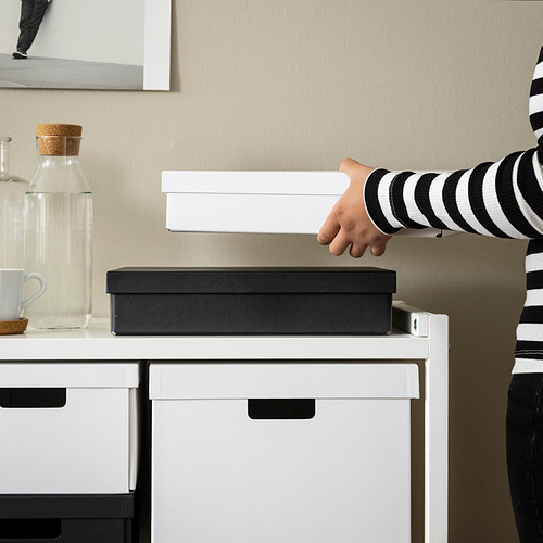 La mano alcanza la caja blanca FJÄDERHARV sobre el escritorio, junto a la caja negra y la jarra, enfatizando el tamaño de las cajas y el diseño moderno.
